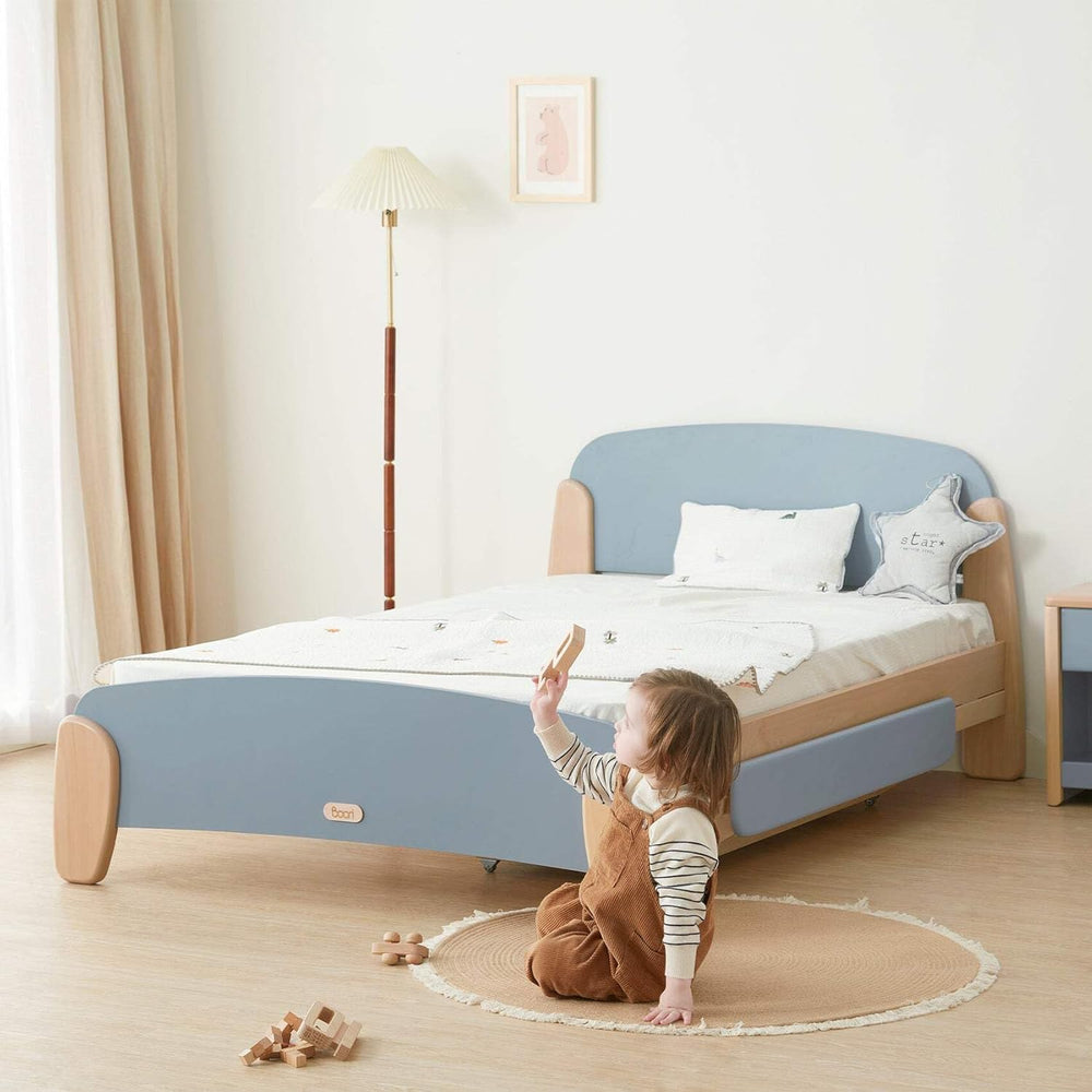 Child playing on the floor next to a blue and wooden bed in a room with a lamp and curtains.