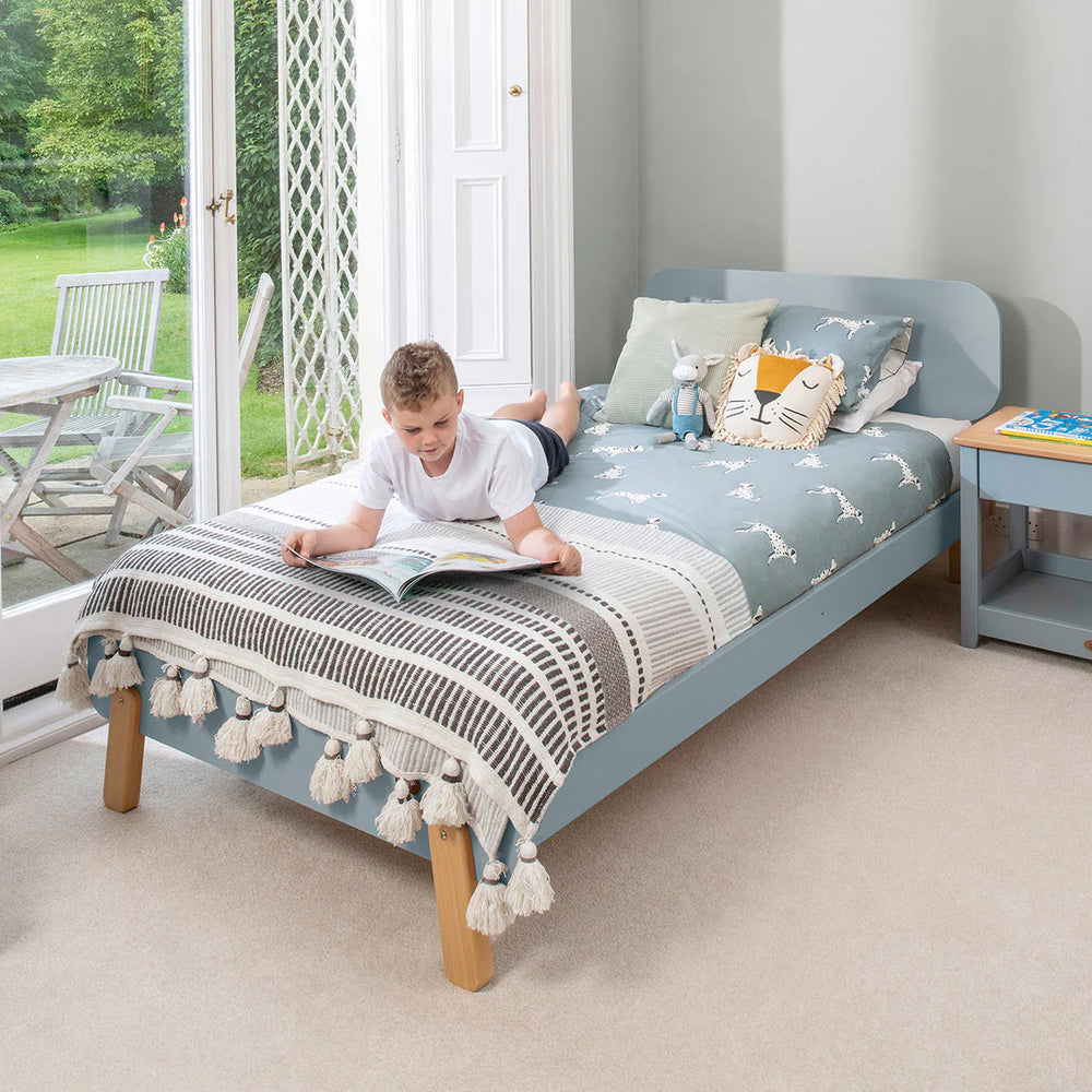 Child reading a book on a blue bed with a striped blanket in a bright room.