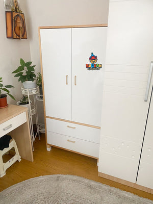 A white wardrobe with a design that includes two doors and a drawer, situated in a room with a desk and a plant to the side.