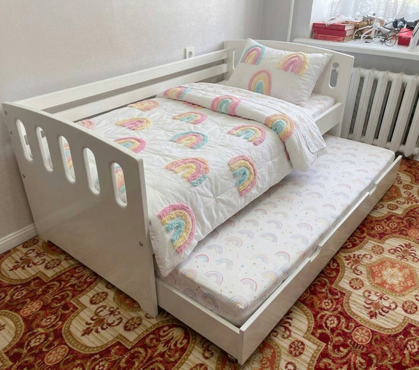 White children's bed with rainbow-patterned bedding on a patterned carpet.